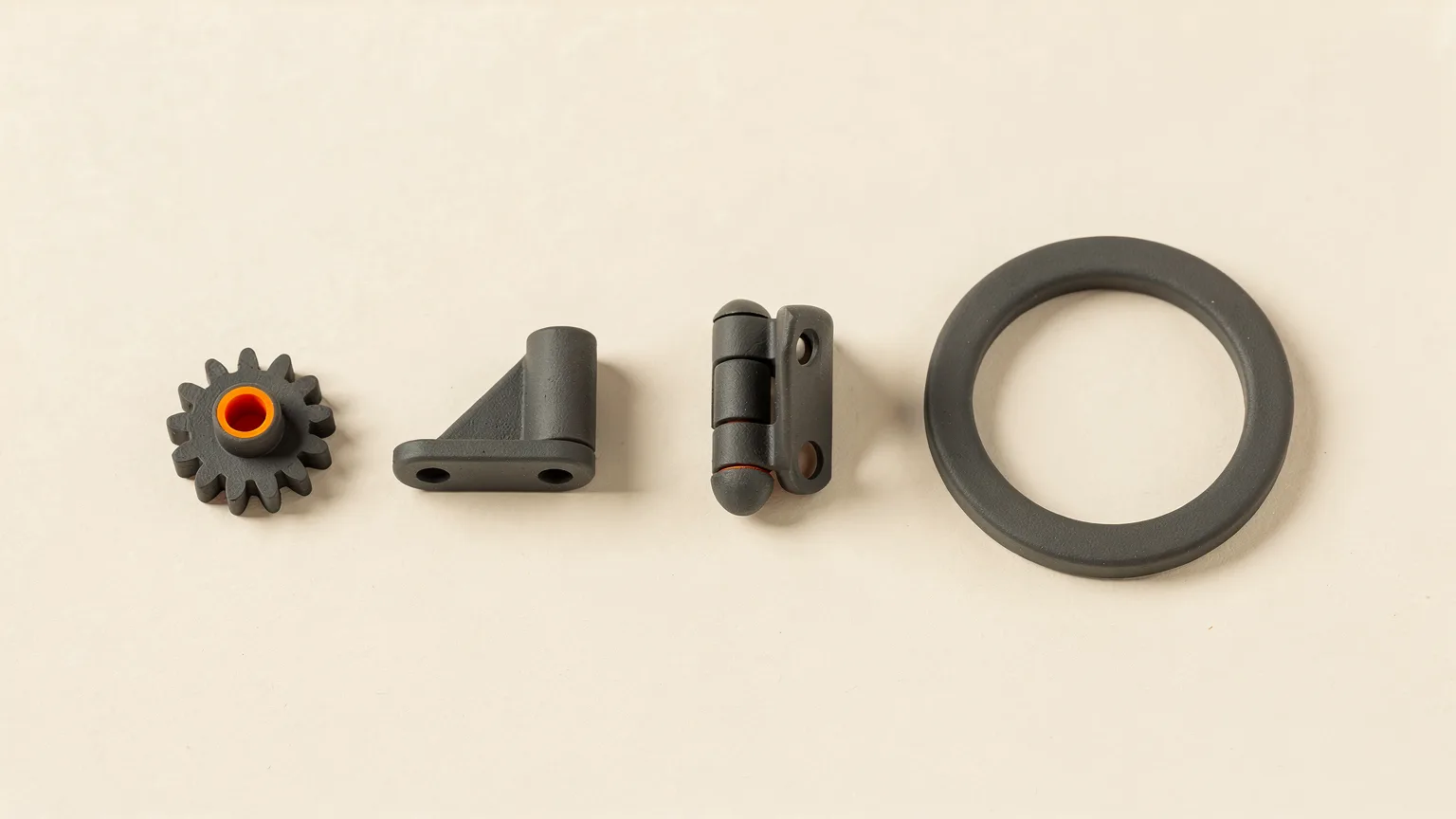 Three dark gray printed parts, a small gear with an orange hub, a bracket and a hinge, beside a flat ring on a cream background