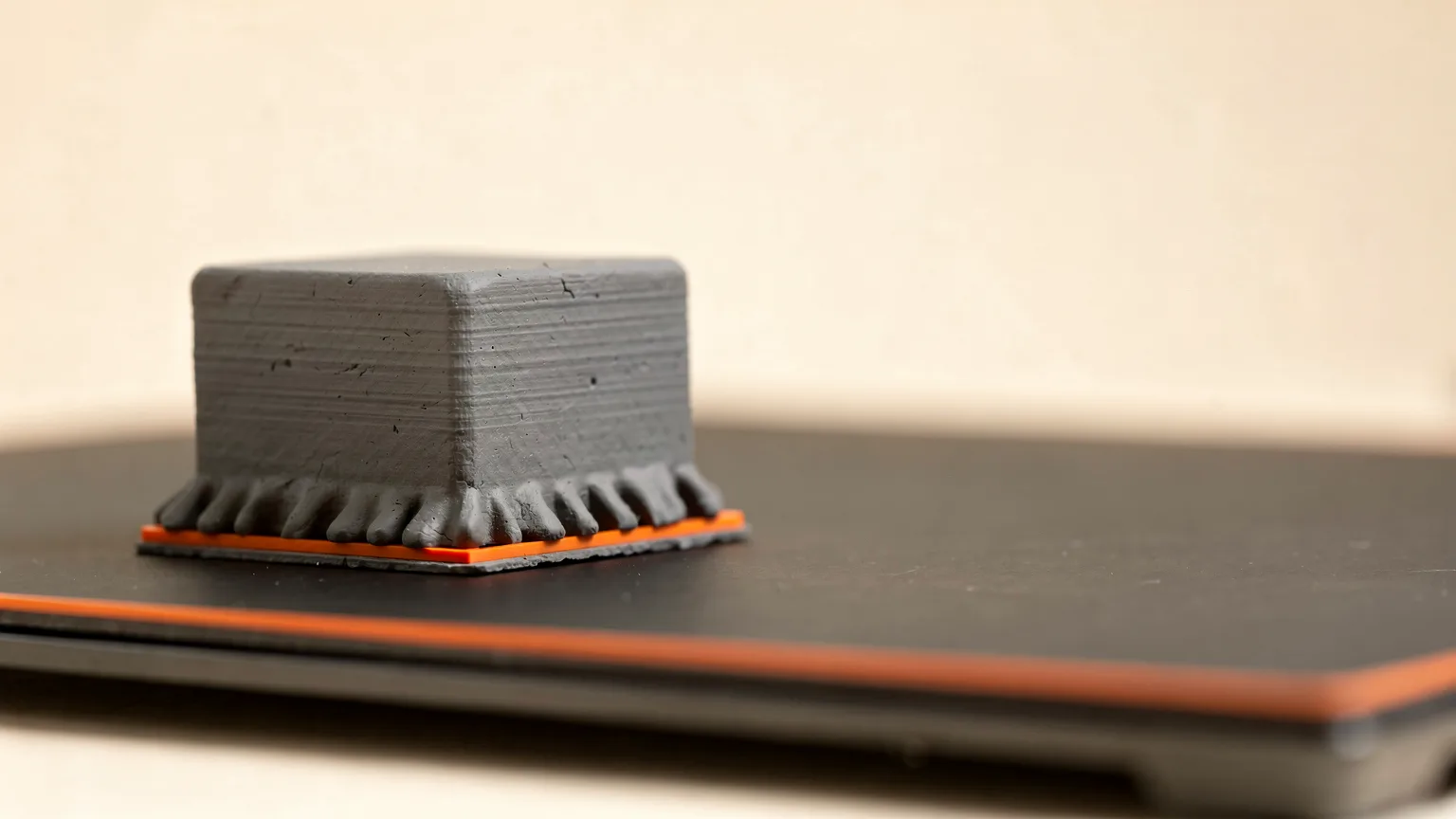 Illustration: a small graphite printed square block with its bottom edge visibly flared outward, next to a dark flat bed, orange first layer line at the base