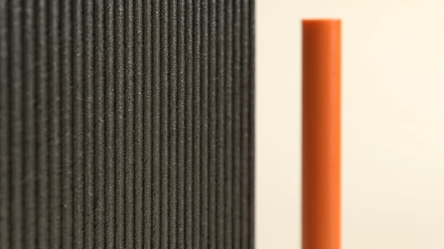 Close view of a graphite printed wall with fine evenly spaced vertical ridges beside a smooth orange pillar