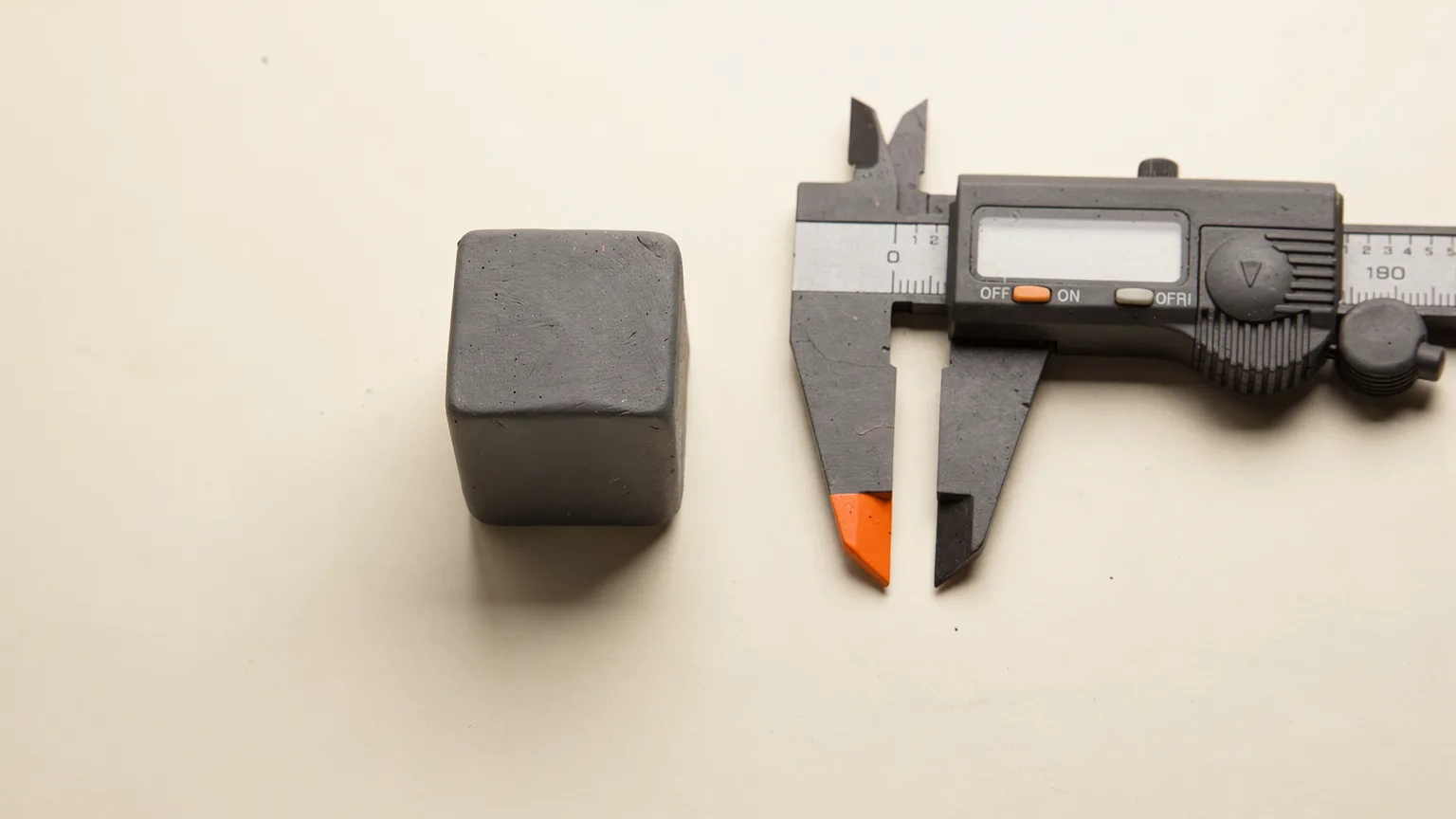 Illustration: a small graphite calibration cube with a pair of precise digital calipers lying beside it, one orange accent on the caliper tip, top-down view