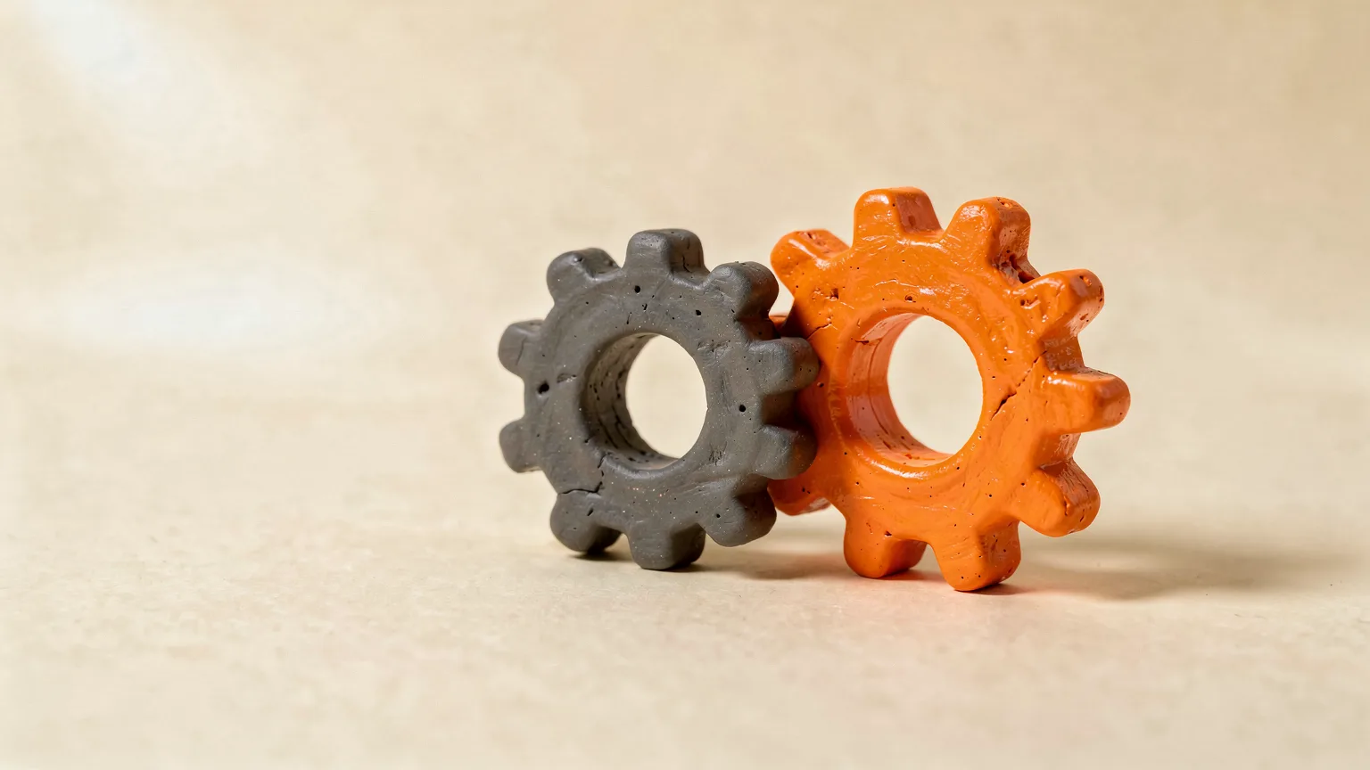 Illustration: two small matching printed gears, one plain graphite and one with a glossy orange finish, sitting on a cream surface