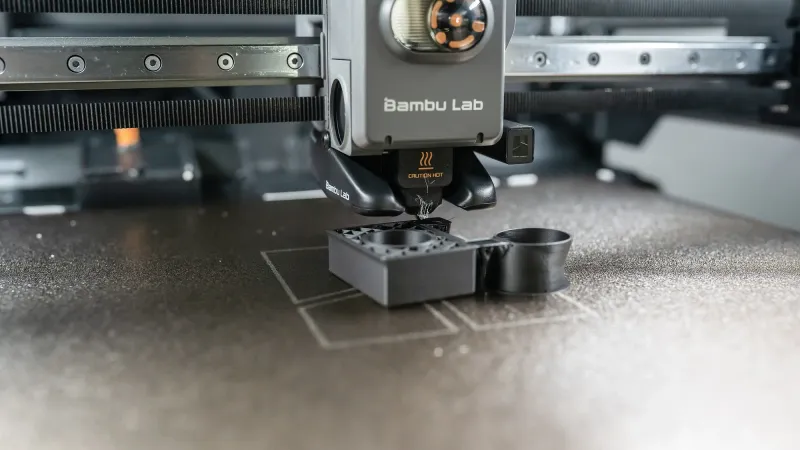 Bambu Lab printer toolhead on its X-axis gantry rail with belt visible above a dark textured build plate