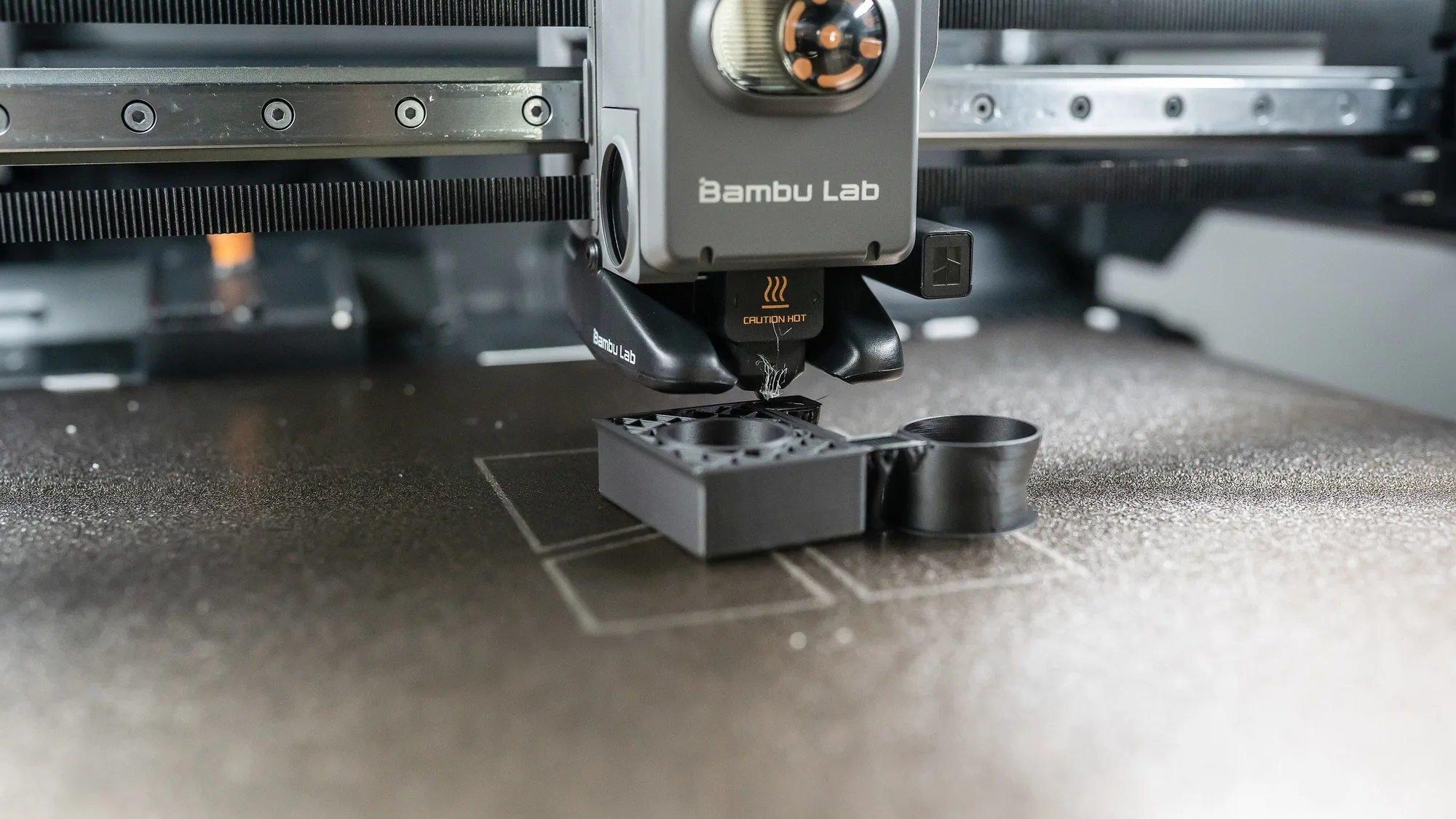 Bambu Lab printer toolhead on its X-axis gantry rail with belt visible above a dark textured build plate