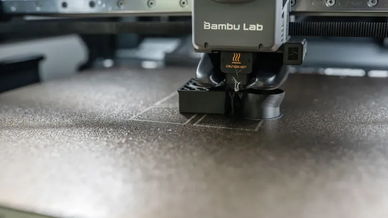 Close-up of a Bambu Lab printer toolhead and nozzle above a dark textured build plate with small grey printed parts on it