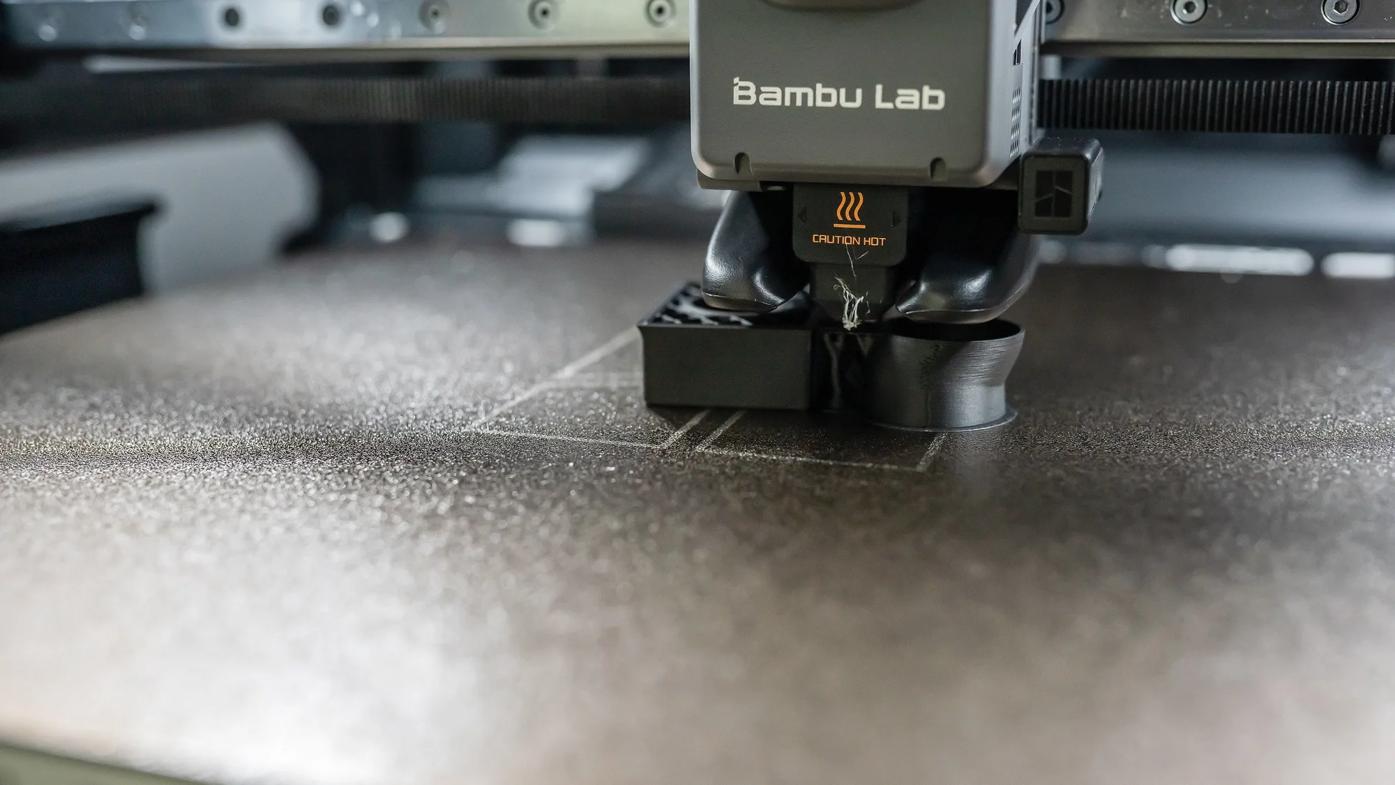 Close-up of a Bambu Lab printer toolhead and nozzle above a dark textured build plate with small grey printed parts on it