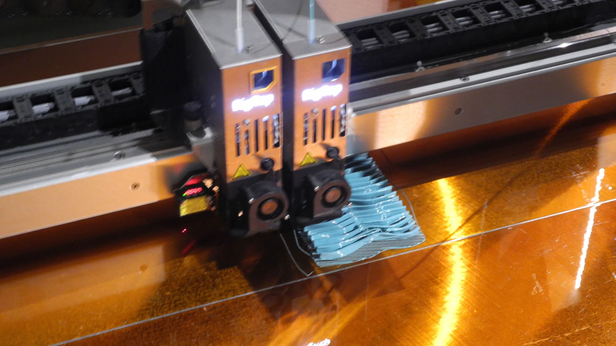 Dual print heads of a large-format 3D printer mid-print, depositing blue plastic layer by layer onto a glossy build plate