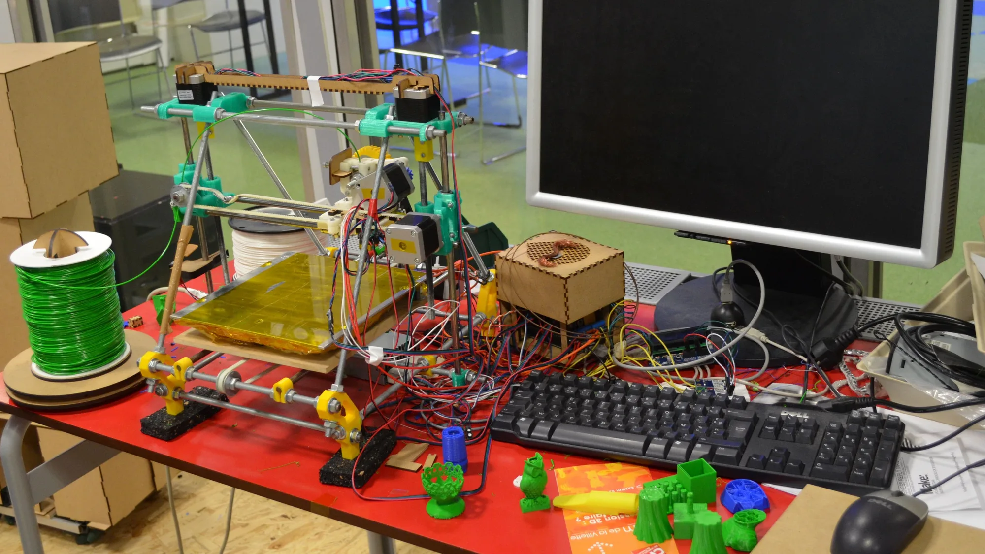 Home-built RepRap-style 3D printer on a red workshop table beside a green filament spool and a group of small green 3D-printed objects