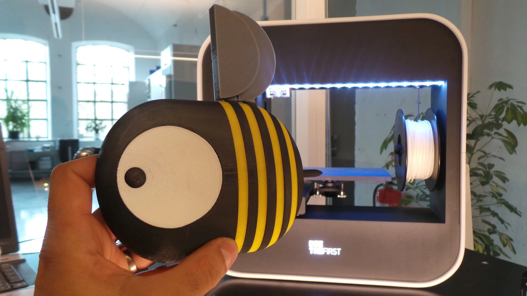Hand holding a bee figure 3D-printed in black, yellow and white parts, in front of a desktop 3D printer with a white filament spool inside