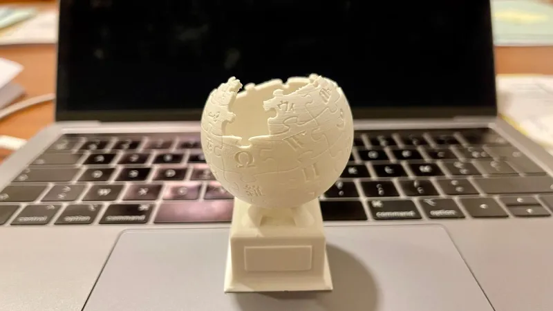 White 3D-printed puzzle-globe trophy standing on the keyboard of a laptop on a desk, showing a finished print up close