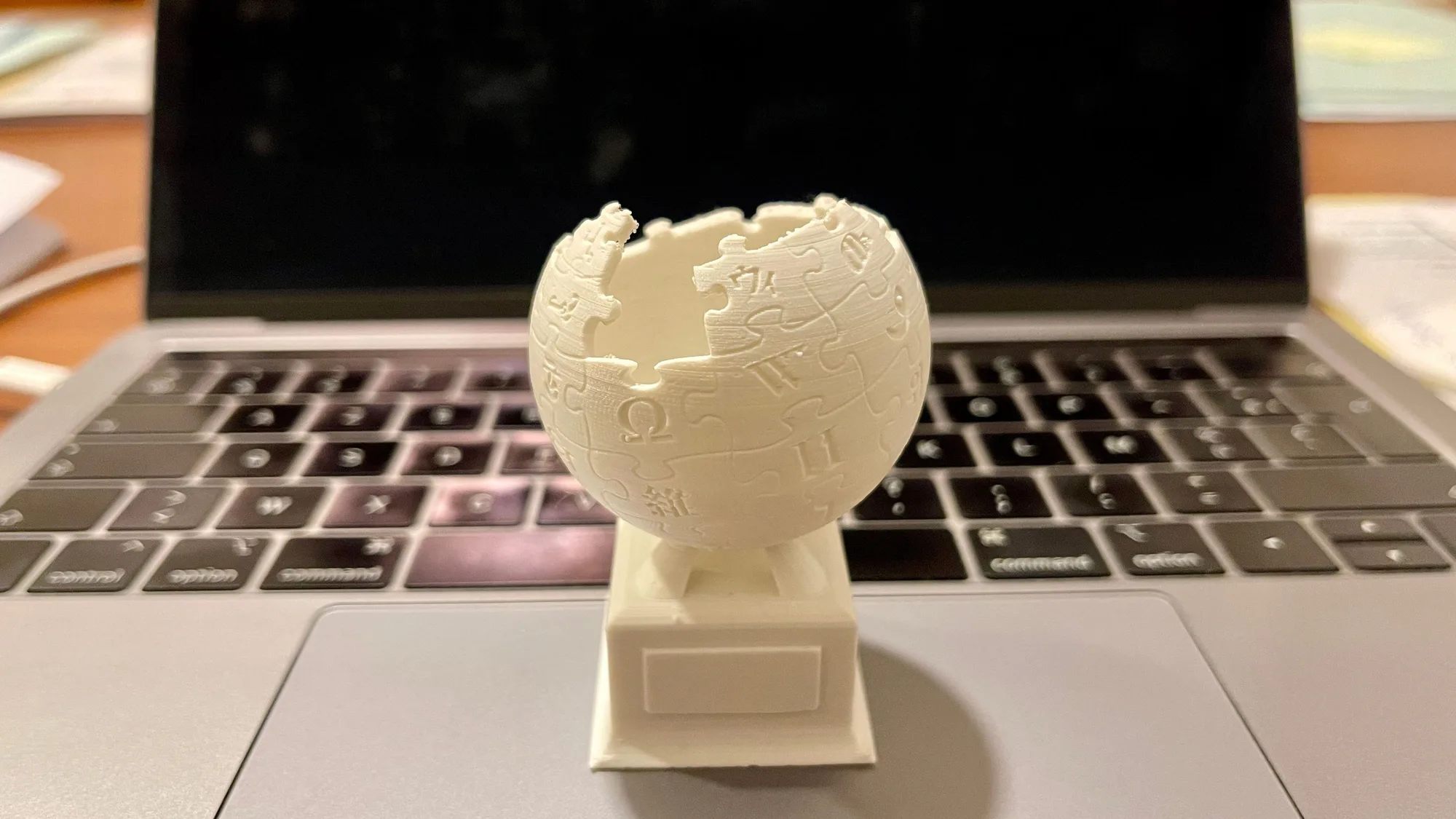White 3D-printed puzzle-globe trophy standing on the keyboard of a laptop on a desk, showing a finished print up close