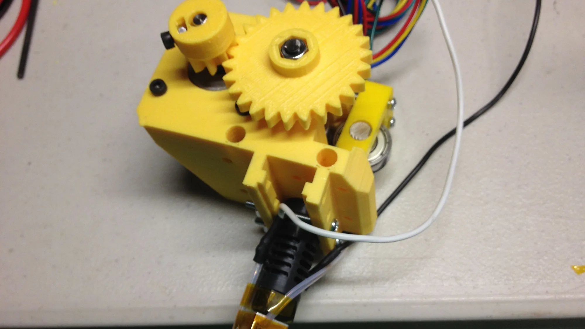 Assembled extruder of a home-built Prusa i3 3D printer on a workbench, with yellow printed gear and body parts, hotend and loose wiring
