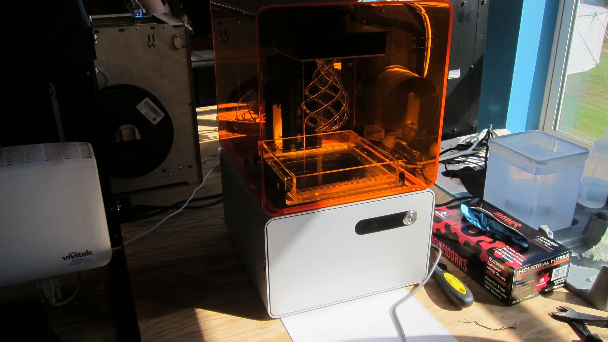 Orange-hooded resin 3D printer printing a spiral lattice on a workbench, with a wooden-framed filament 3D printer and spool behind it