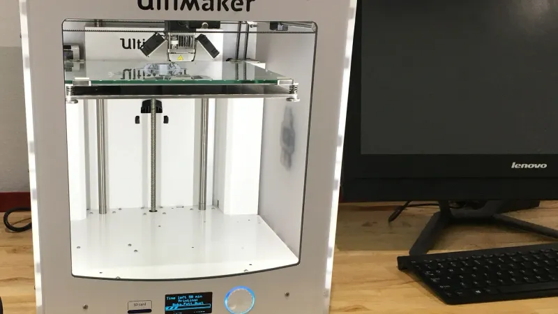 Ultimaker 2 desktop 3D printer printing on a wooden workbench next to a computer monitor and keyboard against a white wall