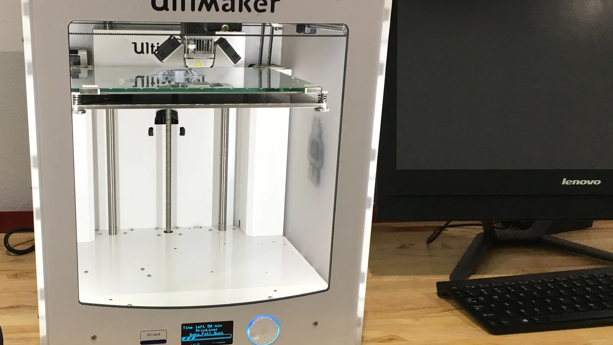 Ultimaker 2 desktop 3D printer printing on a wooden workbench next to a computer monitor and keyboard against a white wall