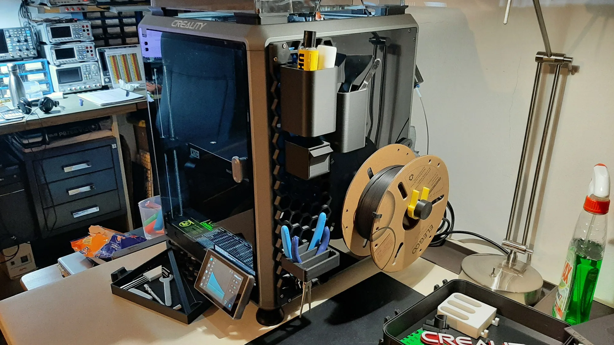 An enclosed Creality K1C 3D printer on a workbench with storage accessories attached and a spool on the side