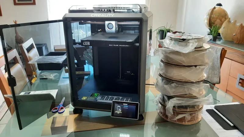 A Creality K1C enclosed 3D printer with its door open beside a stack of five spools of PLA filament on a glass table