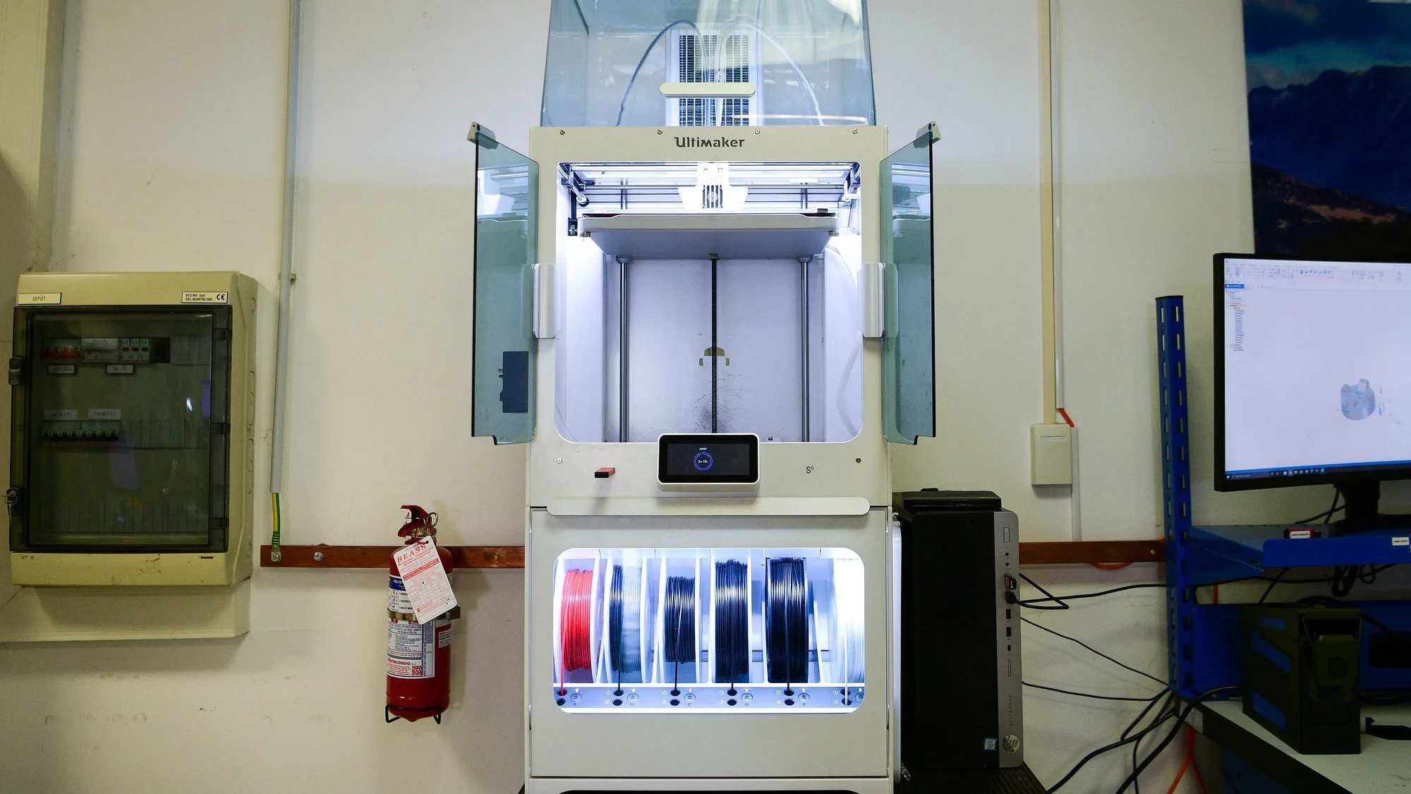 Enclosed Ultimaker 3D printer with open side doors in a workshop, a red fire extinguisher hanging on the wall beside it