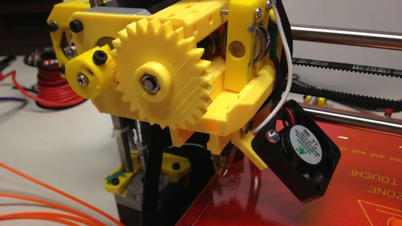 Close-up of a 3D printer extruder with a yellow printed gear and stepper motor above the hot end, orange filament feeding in