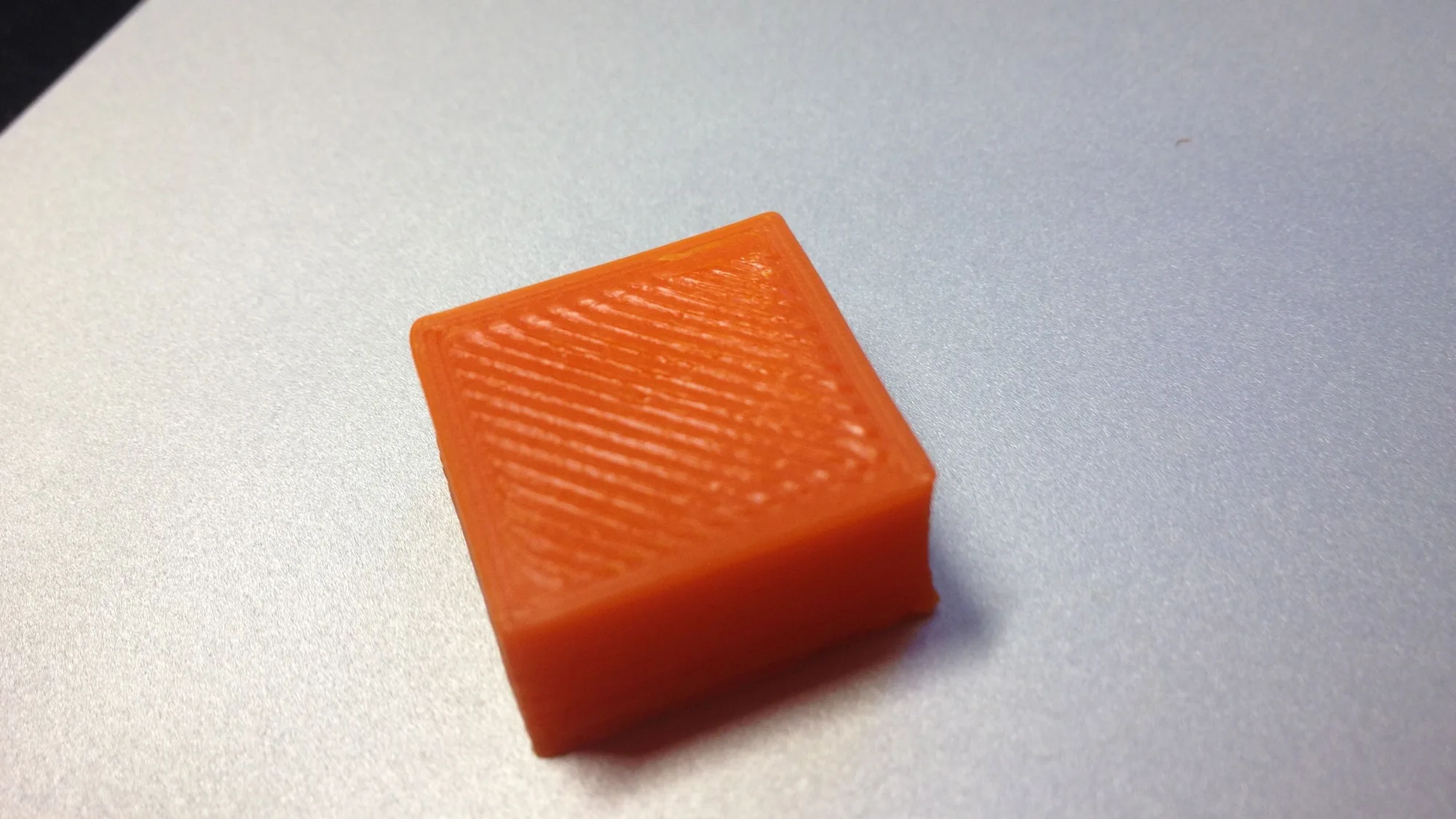 A small orange 3D printed test cube resting on a laptop, showing the top infill lines and layered sides