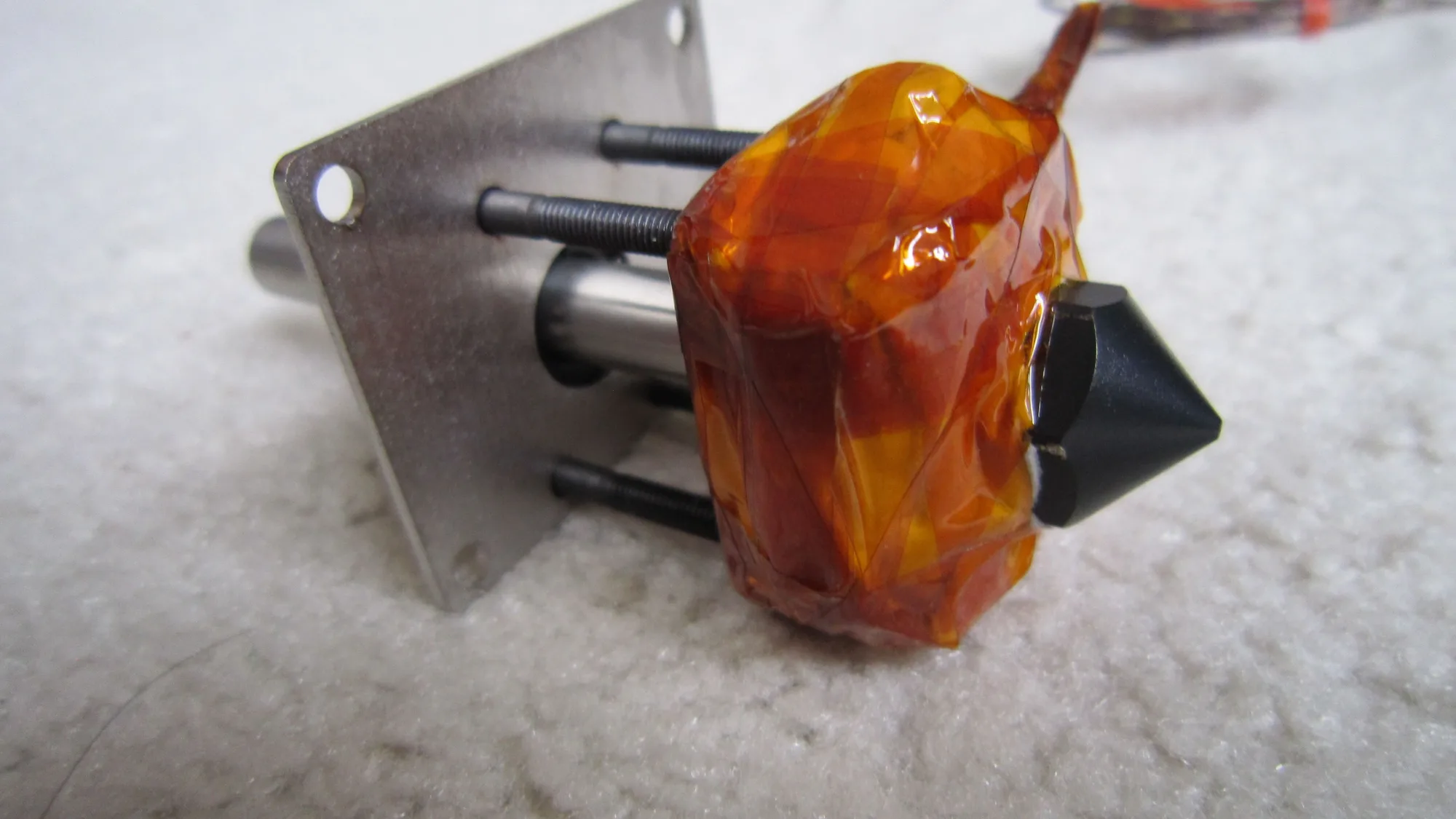 A hotend wrapped in orange Kapton tape with a black cone-shaped nozzle on the right and a metal mounting plate on the left