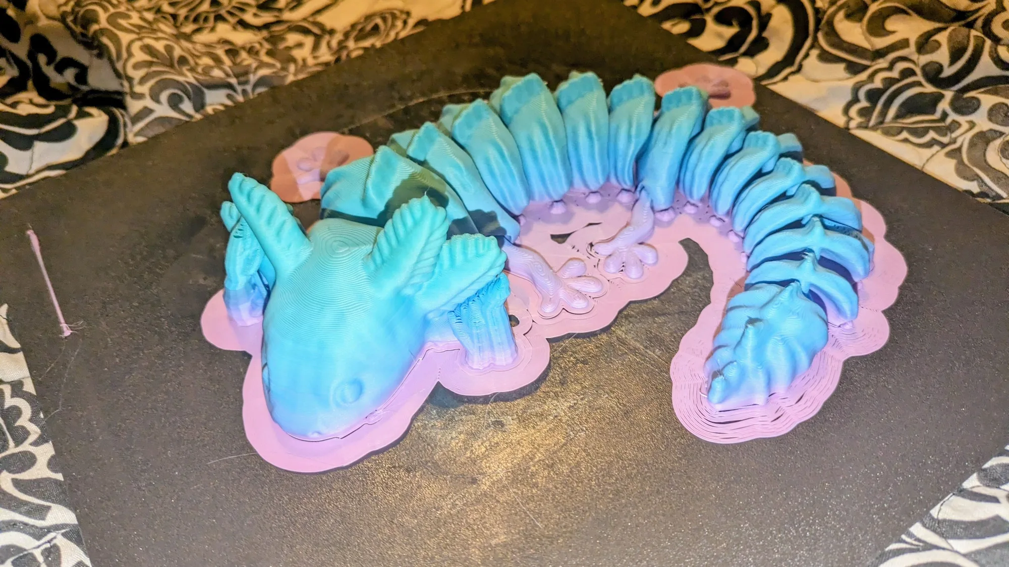 A finished two-colour 3D printed axolotl with a pink brim and base layers still sitting on a dark build plate