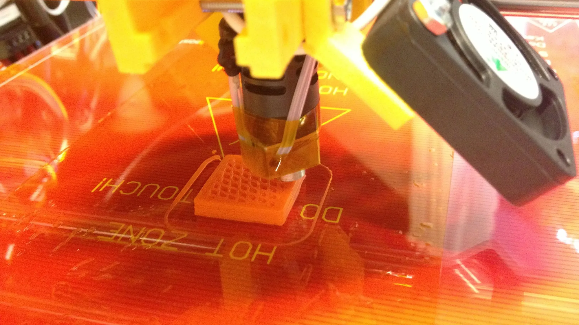 A 3D printer nozzle building a small orange block with a visible grid infill pattern on a heated bed