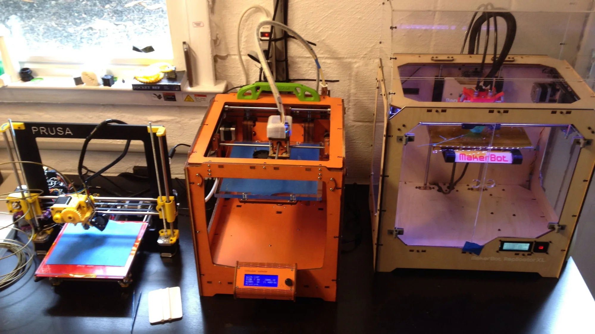 Three hobbyist 3D printers side by side on a workbench: a Prusa i3, an orange open-frame printer with an LCD controller and…