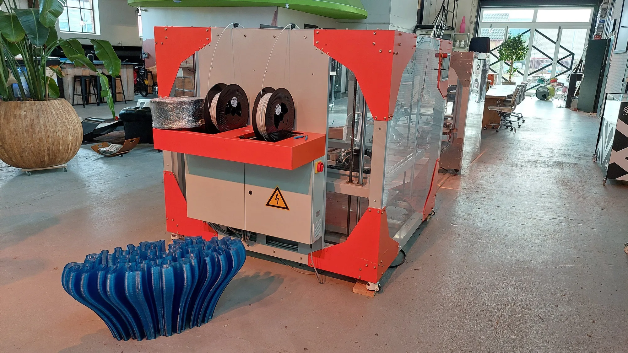 Large-format 3D printer with two filament spools mounted on top, standing in a makerspace beside a big blue printed vase-shaped part