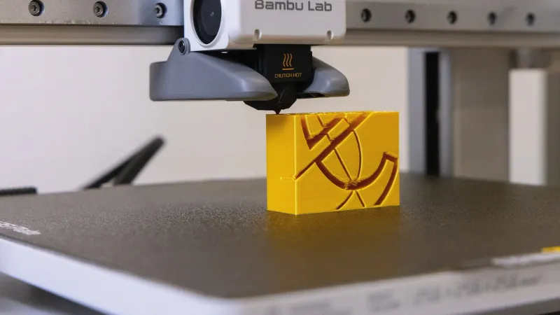 Bambu Lab toolhead with its nozzle just above a yellow printed block on the textured print bed of a desktop 3D printer