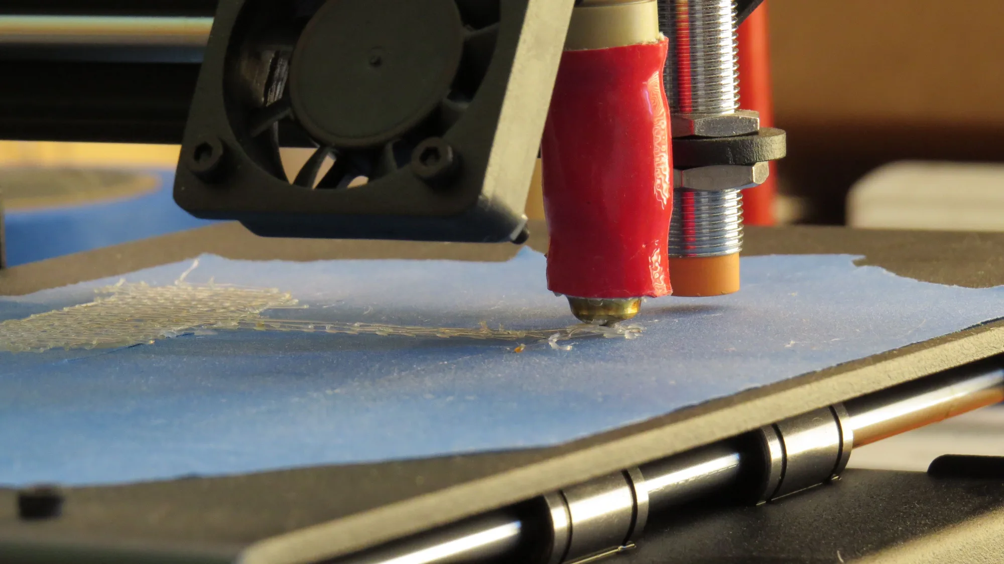 A 3D printer nozzle laying down the first layer on a blue taped print bed