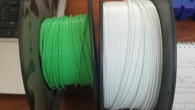 Two spools of PLA filament, one green and one white, side by side on a wooden desk