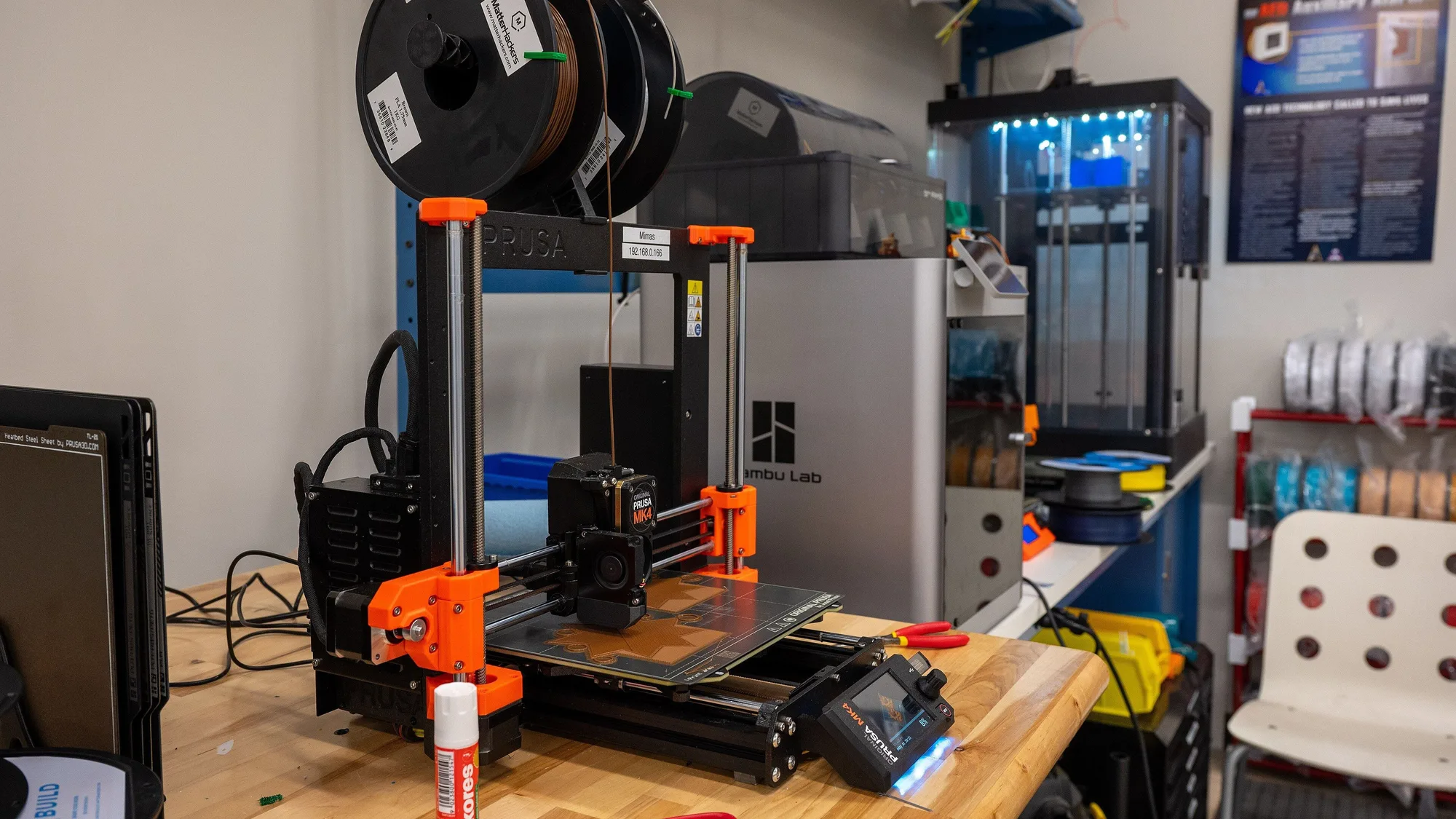 Prusa i3-style 3D printer on a wooden workbench with filament spools above it, side cutters, scissors and a glue stick beside it