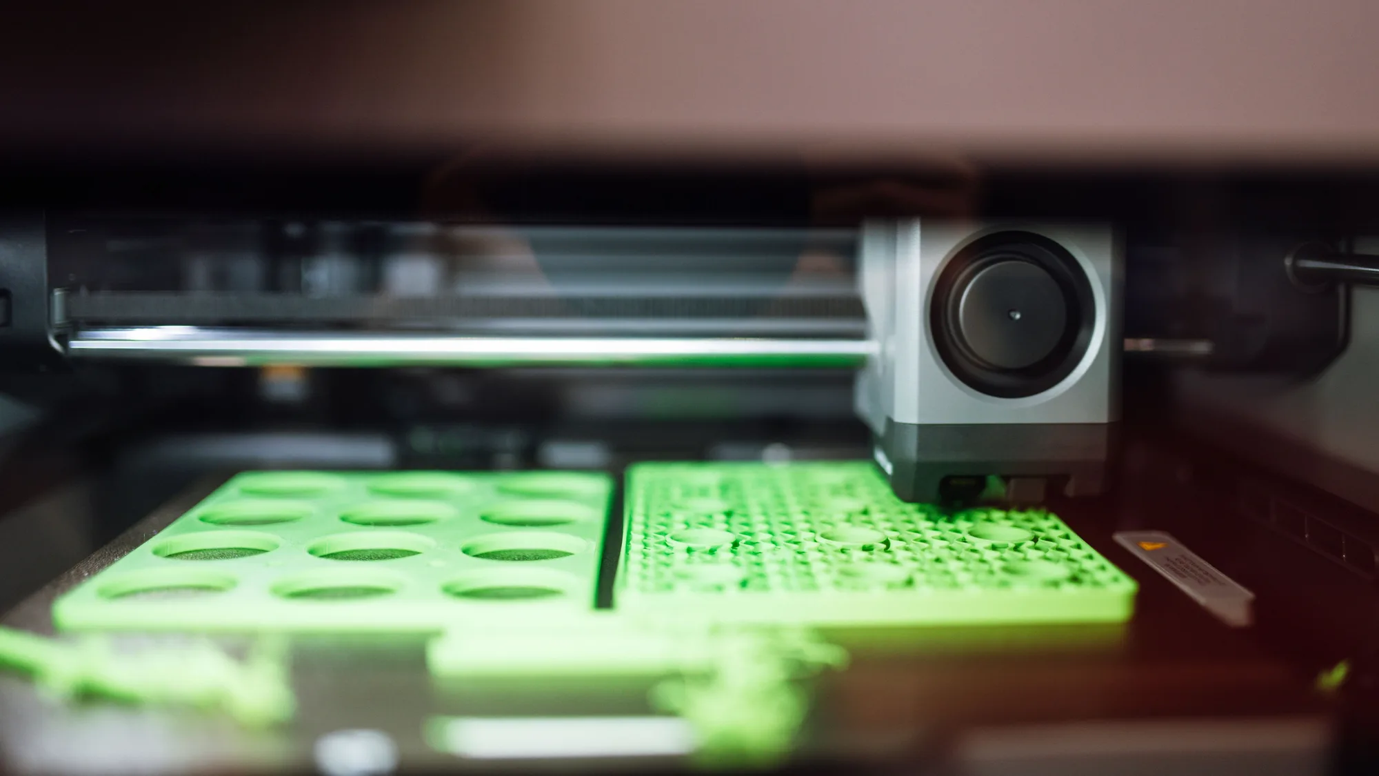 Close-up of a 3D printer toolhead printing flat green plates on the print bed