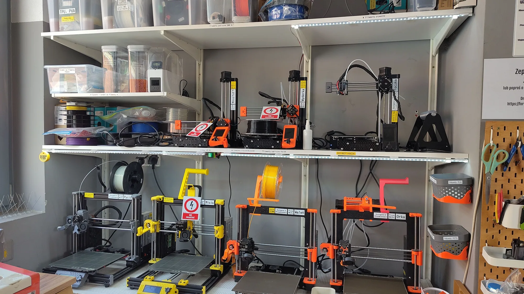 A workshop shelf with several 3D printers lined up and storage boxes above