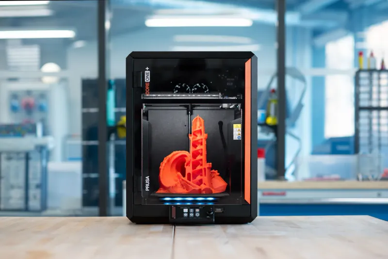 Original Prusa CORE One+ enclosed 3D printer on a workbench with an orange lighthouse model printed inside, blue-lit workshop