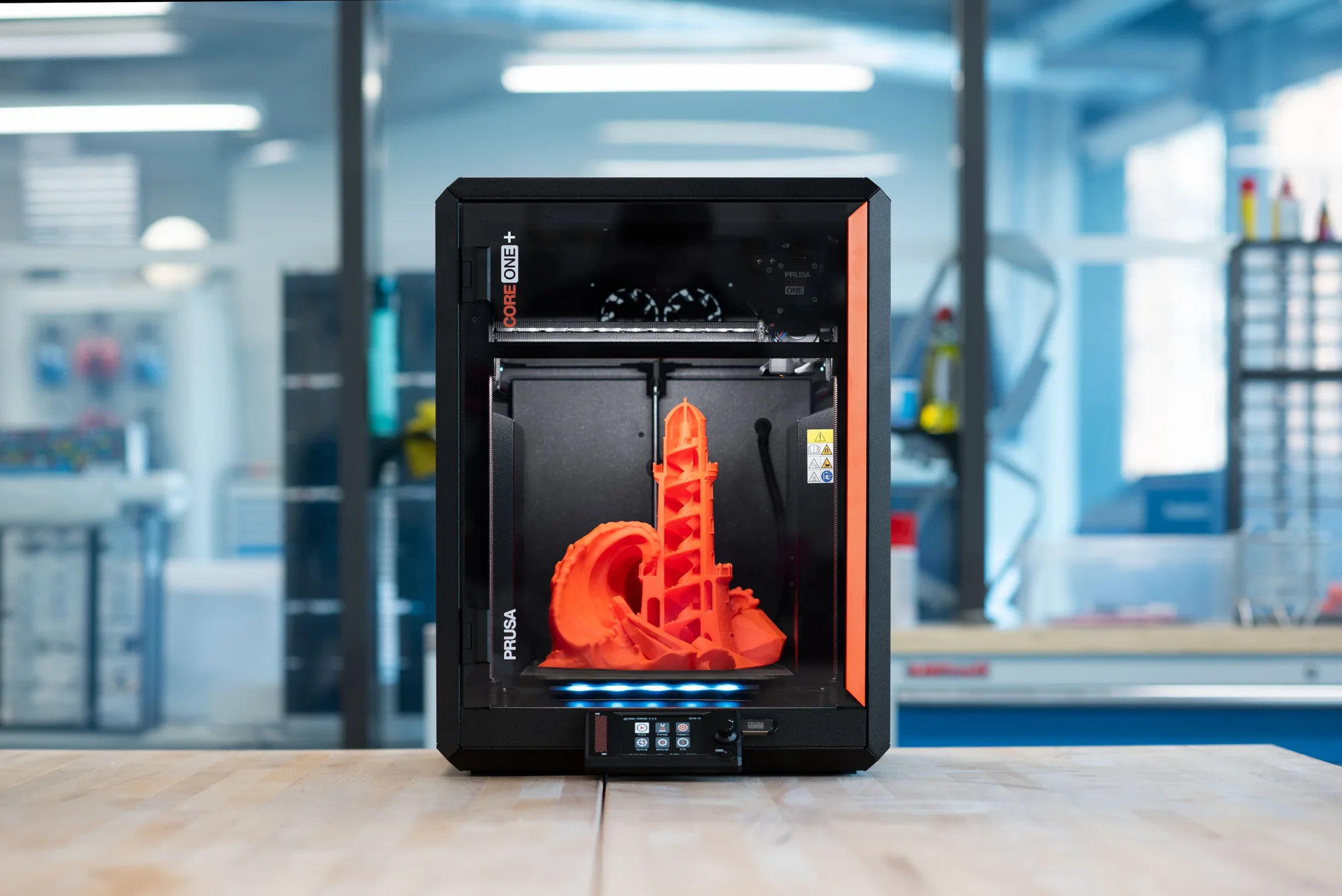Original Prusa CORE One+ enclosed 3D printer on a workbench with an orange lighthouse model printed inside, in a blue-lit workshop